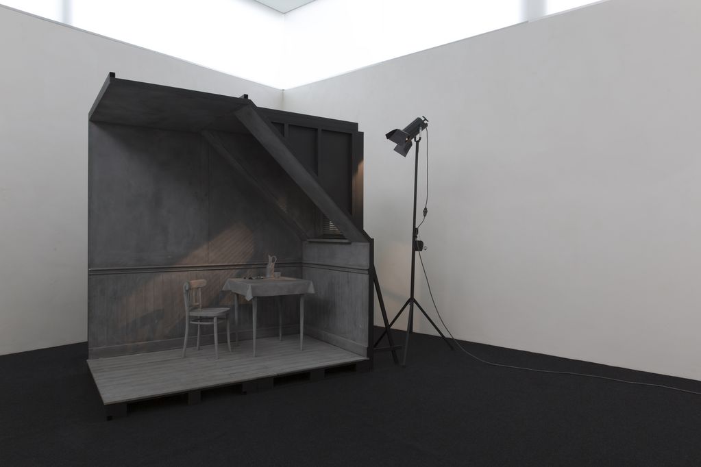 Auf der Installationsansicht ist ein graues, maßstabgetreues Modell einer Zimmerecke mit Stuhl, gedecktem Tisch sowie einem Fenster zu sehen, durch das künstliches Licht von einer außenstehenden Lampe fällt.