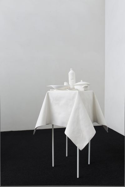 A white plaster table with long, thin table legs stands on a black ground in the lower right center of the image. The table itself is covered with a tablecloth that casts even wrinkles over the edges of the table, on top of which are a bottle, a small plate, a pot with a lid and a large plate. The complete model is made of white plaster, the background is painted white. 