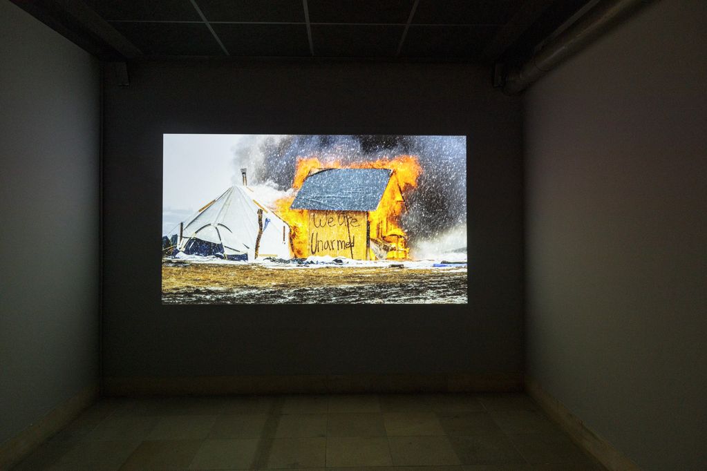 Die Aufnahme eines weißen Zeltes neben einer brennenden Hütte mit der Schrift "We are unarmed" wird auf eine Wand projieziert. Cyrill Lachauer, Sammlung Goetz, München