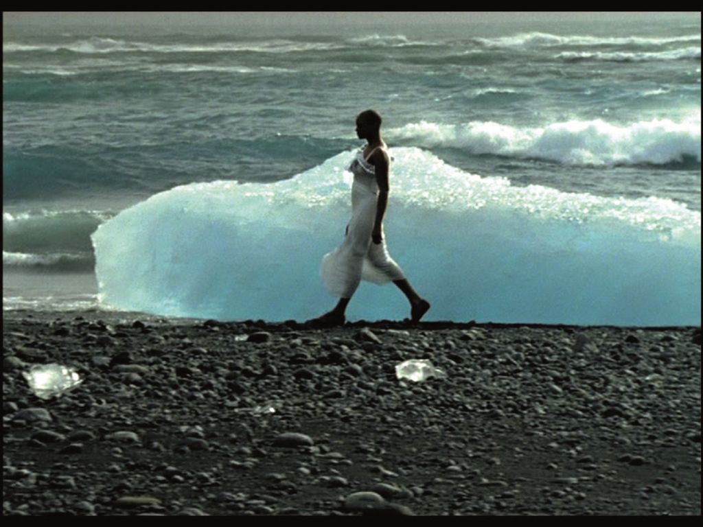 This film still shows a colored, walking woman in profile, in front of an ice floe, behind it the waves break on the beach.