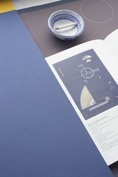 Diese Fotografie zeigt ein aufgeschlagenes Heft mit einer Farbschema-Zeichnung Kandinskys, sowie ein kleine weiße Schale mit blauer Bemalung und einem weißen Holzbuntstift darin. Links daneben liegt Tonpapier in einem warmen, hellen Blauton.