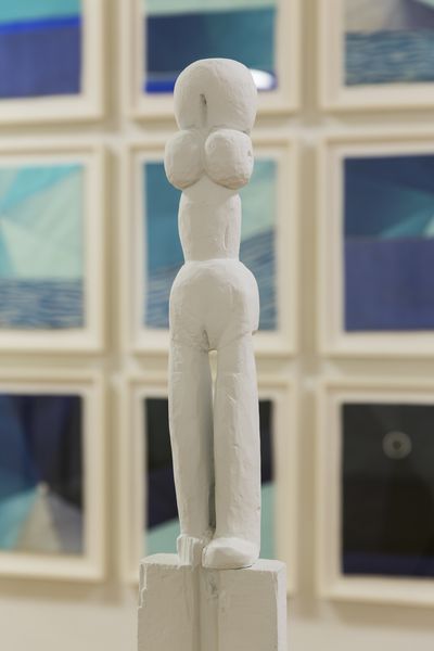 Diese Installationsansicht zeigt eine weiß bemalte, weibliche und langgezogene Skulptur vor einem Hintergrund einer mehrteiligen, zweidimensionalen und bläulichen Arbeit aus Stoff.