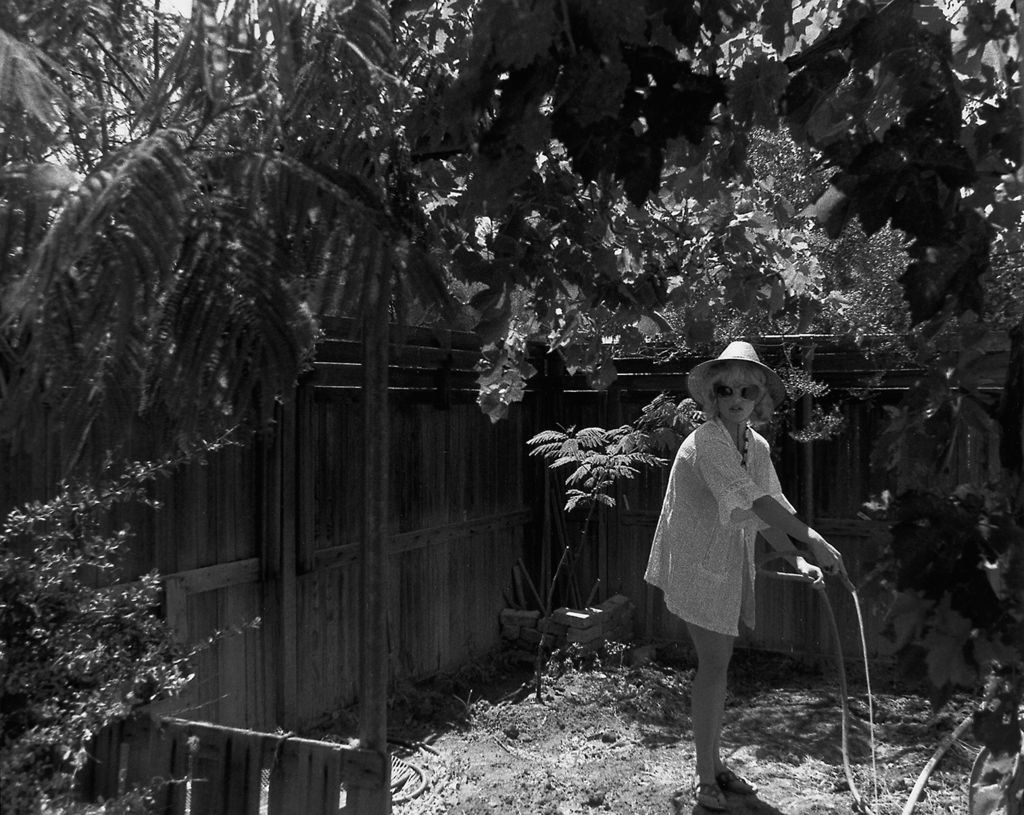 Black and white photograph of a woman dressed in a bright tunic and sun hat, looking in the direction of the viewer as she waters her garden with a garden hose. Cindy Sherman, Sammlung Goetz Munich