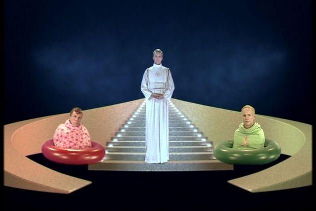 Veraltetes Science-Fiction Szenario mit einer weiß gekleideten Gestalt auf einer Treppe links und rechts zwei halbe Gestalten in einem Gummiring 