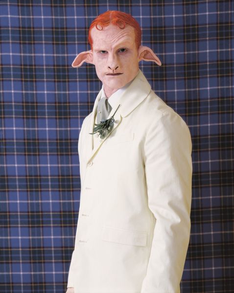 In this photograph you can see a Satyr-like creature, with bright red hair, a white suit with a lapel plant and a neckpiece. It stands in front of a fabric of dark blue tartan pattern.