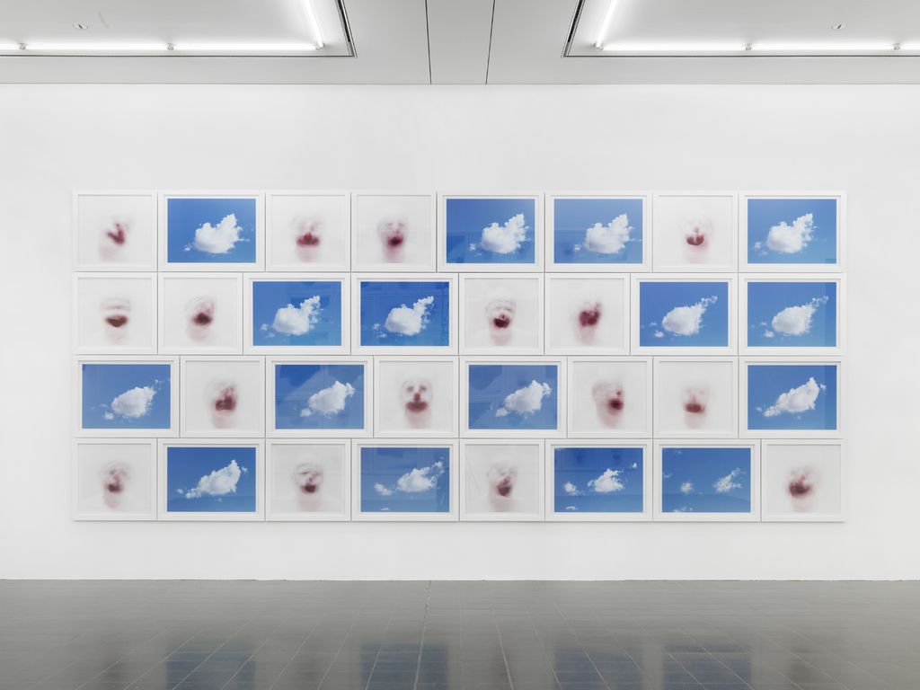 This installation photo shows an arrangement of photographs, alternating between clouds in a rich, blue sky and blurred clown faces.