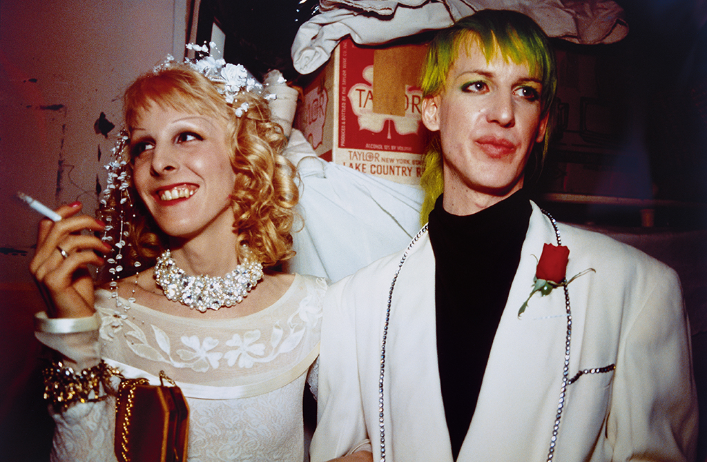 The photograph shows a bridal couple, the section is at breast height. The bride has a cigarette in her hand and is laughing, while the groom is wearing a white jacket and shows only an implied smile. Both wear make-up.