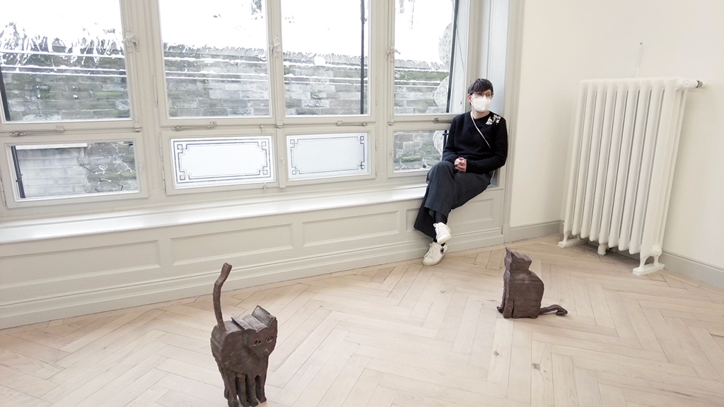 Photography of the artist with mouth-nose protection. In front of her on the floor are two bronze cat sculptures. Sammlung Goetz, Munich