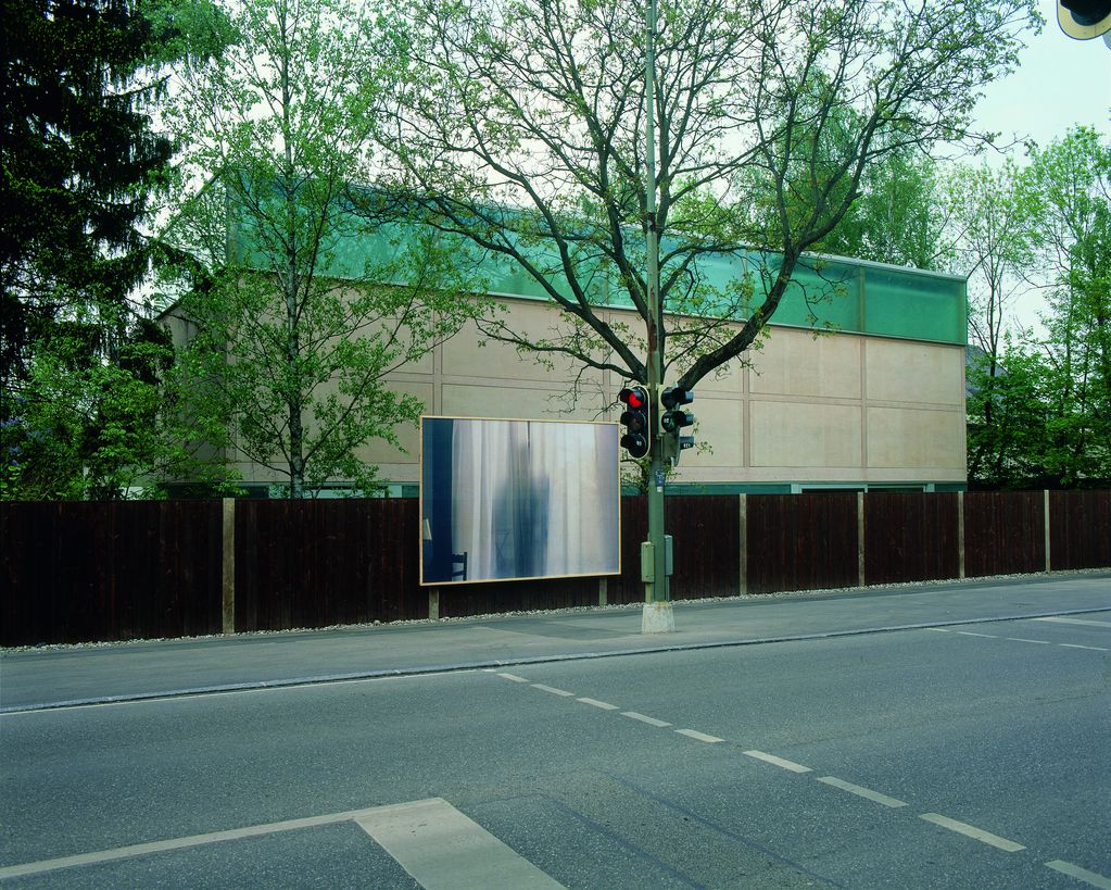 Das Bild zeigt eine Außenaufnahme des Ausstellungsgebäudes von der Straße aus gesehen. Am durchgängigen, dunkelbraunen Zaun ist eine große Werbetafel angebracht, die eine fotografische Arbeit von Felix Gonzalez-Torres zeigt. Darauf zu sehen ist ein weißer, durchscheinender Vorhang mit einer Person dahinter, die einen leichten Schatten wirft.