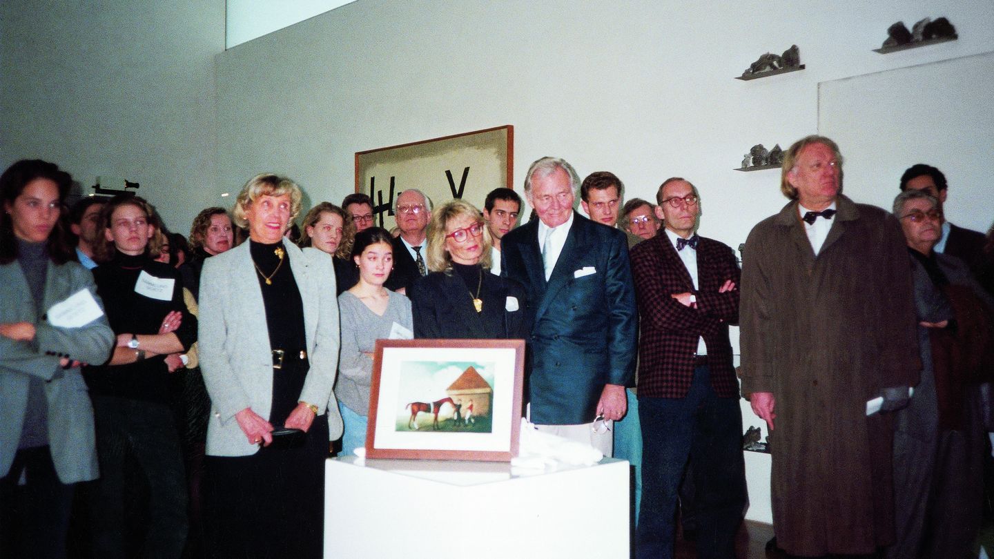 The photograph shows the visitors of the exhibition in the space, crowded together. In the foreground is a photograph on a white pedestal, behind the audience is a work by Jannis Kounellis on the wall.