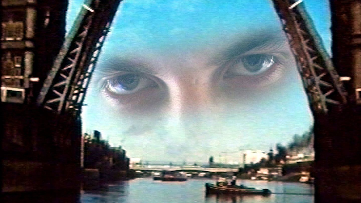 Dieser Filmstill zeigt zwei übereinander gelagerte Aufnahmen von der London Tower Bridge, welche ihre Klappen geöffnet hat, um größere Schiffe durchzuschleusen. Im Hintergrund befindet sich tatsächlich ein kleinerer Dampfer, aus dem schwarzer Rauch herausdringt. In dem oberen Bildteil befindet sich wiederum ein Augenpaar, das intensiv in die Kamera blickt und von den geöffneten Schleusen flankiert wird.