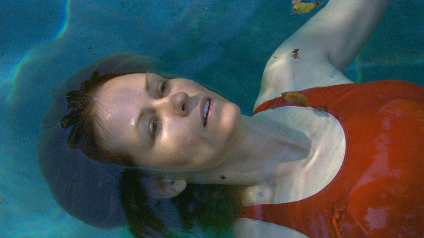 Video Still einer Frau, die in dem türkisblauen Wasser eines Swimming Pools zu schweben scheint. Sie hat weiße Haut, brünettes Haar und feine Gesichtszüge und trägt einen tomatenroten Badeanzug. Teresa Hubbard/Alexander Birchler, Sammlung Goetz München