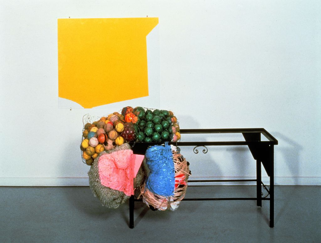 This installation shot contains a two to three dimensional work by Jessica Stockholder. It consists of an acrylic painting in yellow on glass, a black iron frame of a small table and colorful balls in rabbit wire, as well as wrapped sheep wool and other wrapped textiles tied to the table frame. 