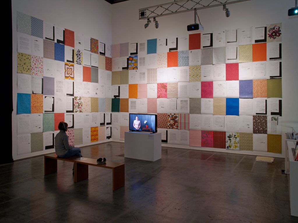 Installation view consisting of a paper installation covering the walls being seen and a 1-channel video installation with monitor and a visitor sitting on a bench with headphones on watching the video. Andrea Bowers, Sammlung Goetz Munich