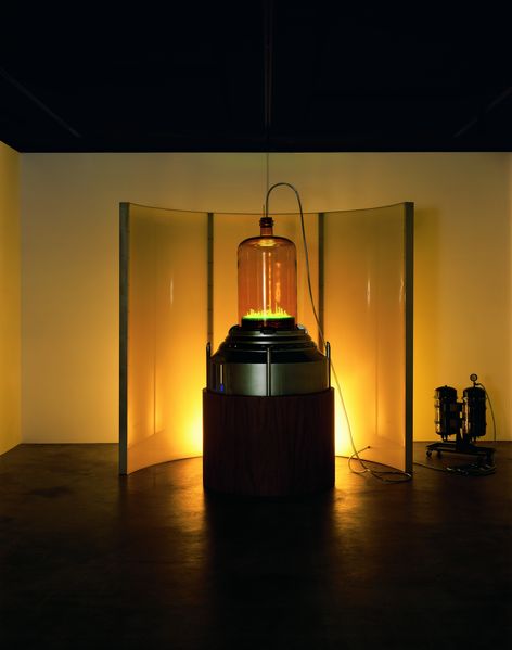 Diese Fotografie zeigt die Ausstellungsansicht der Arbeit "Kandor 7" von Mike Kelley. Im Hintergrund wird ein halbrund gebogener Paravent von einer gelben Lichtquelle beleuchtet, davor steht eine futuristische Skulptur, die eine leuchtende Modellstadt unter einer Glashaube beinhaltet. Mike Kelley, Sammlung Goetz München